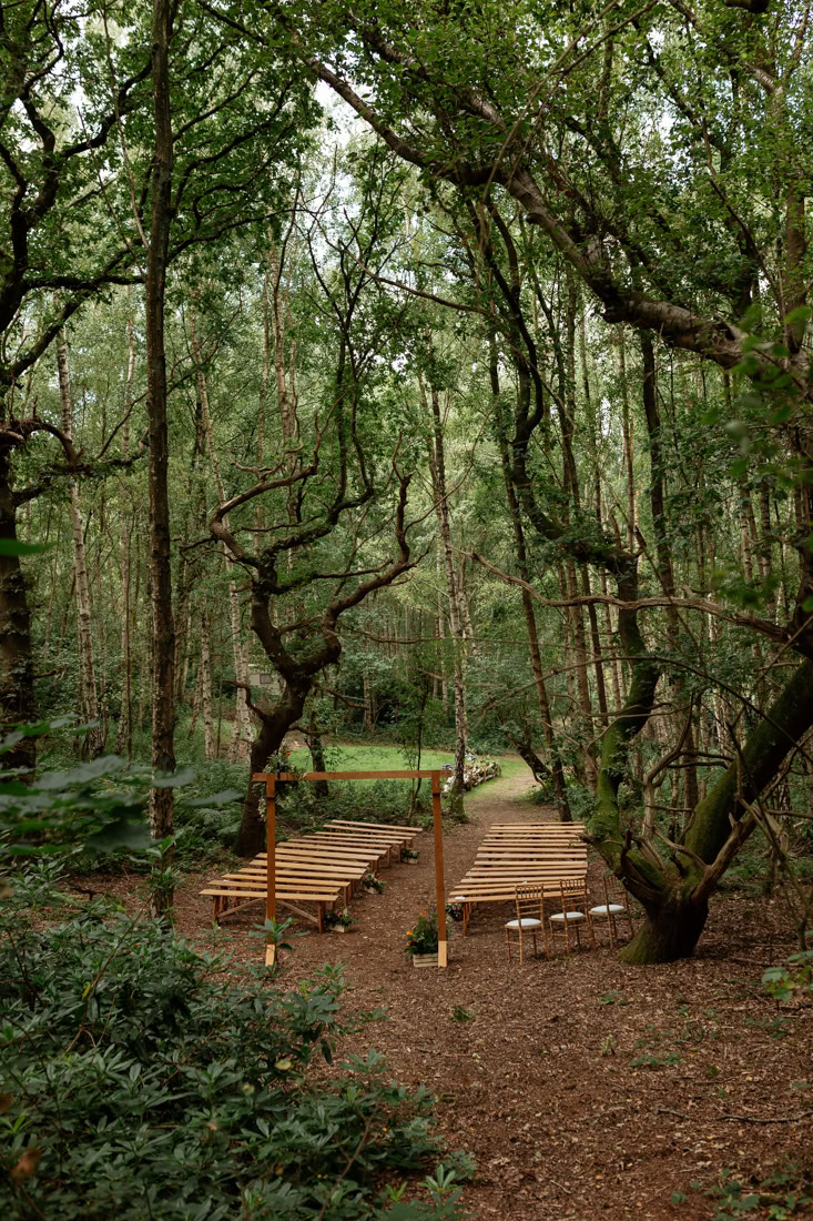 Sherwood Glade wedding Forest ceremony setup with wooden benches lining a path leading to a rustic arch among tall trees.