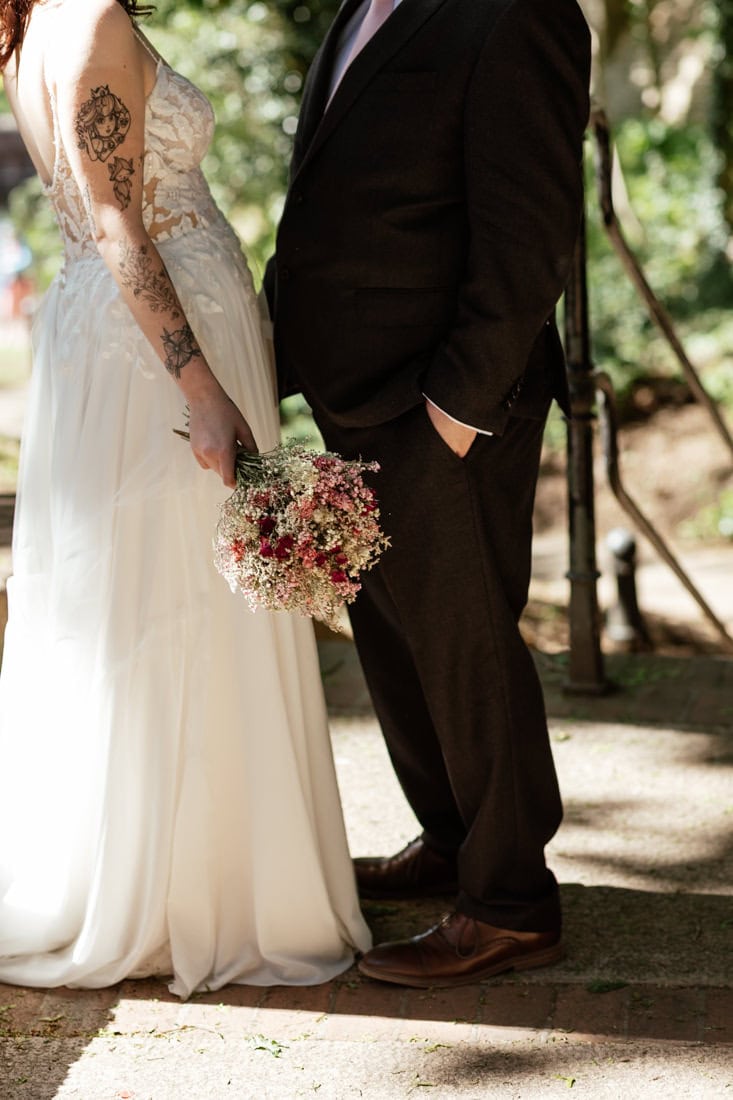 Couple standing outdoors, one in a lace wedding dress holding a dried flower bouquet and the other in a dark suit.