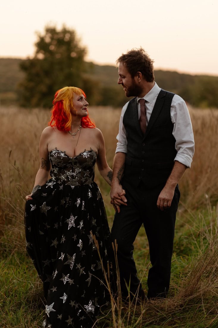 Couple smiling and walking through long grass in wedding attire, she's wearing a black dress with stars, he's in a waistcoat 