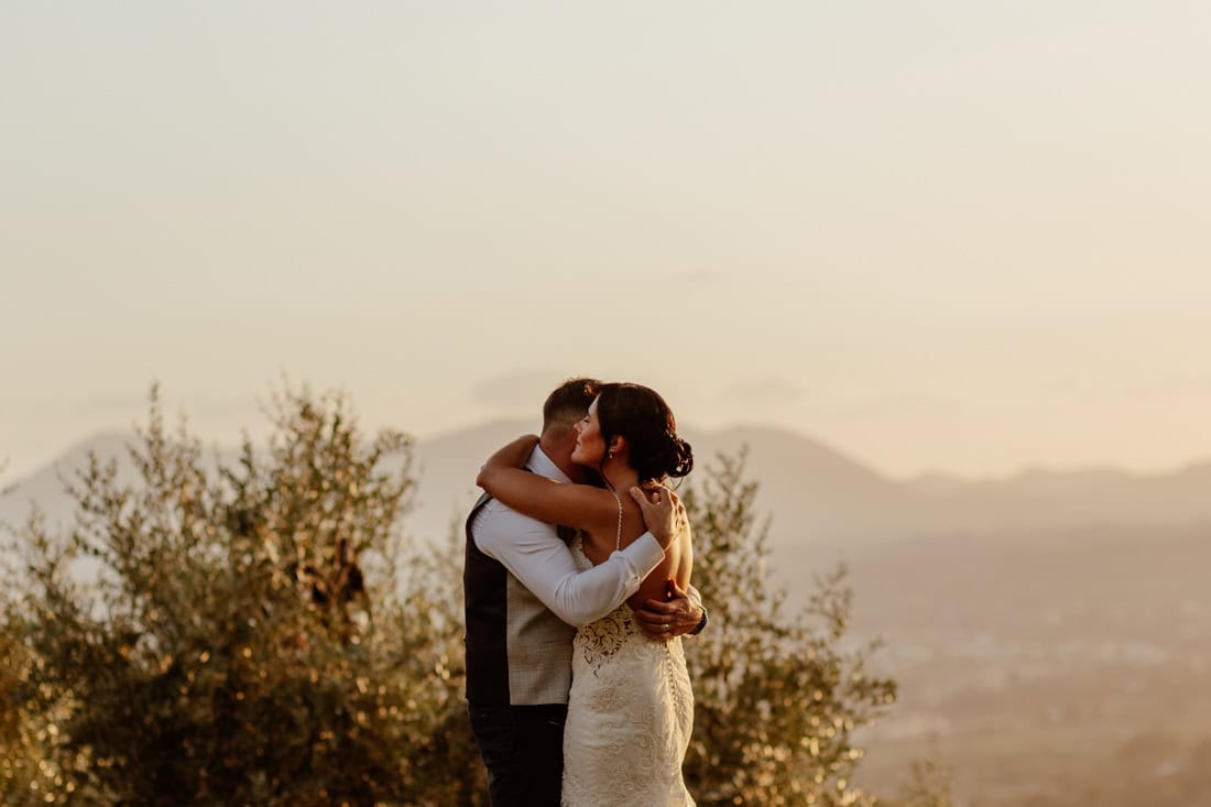 Newlyweds embracing outdoors during a golden sunset, surrounded by trees and distant mountains at this intimate Tuscany wedding