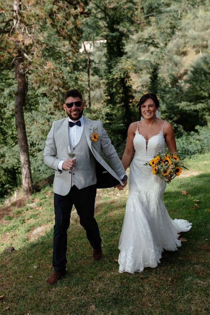 Bride in white lace wedding dress holding a bouquet of sunflowers, walking outdoors with groom in grey suit at this intimate Tuscany wedding.