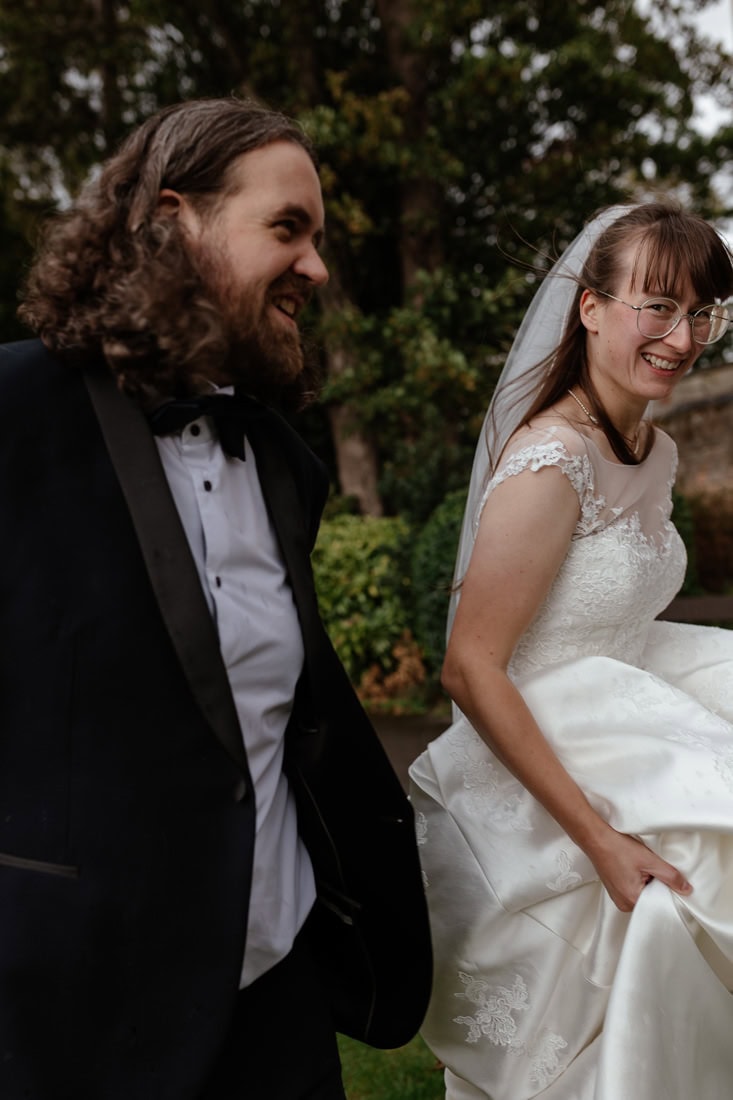 Bride and groom walking through Arnot Hill park smiling, she is carrying her dress and glancing happily at the camera