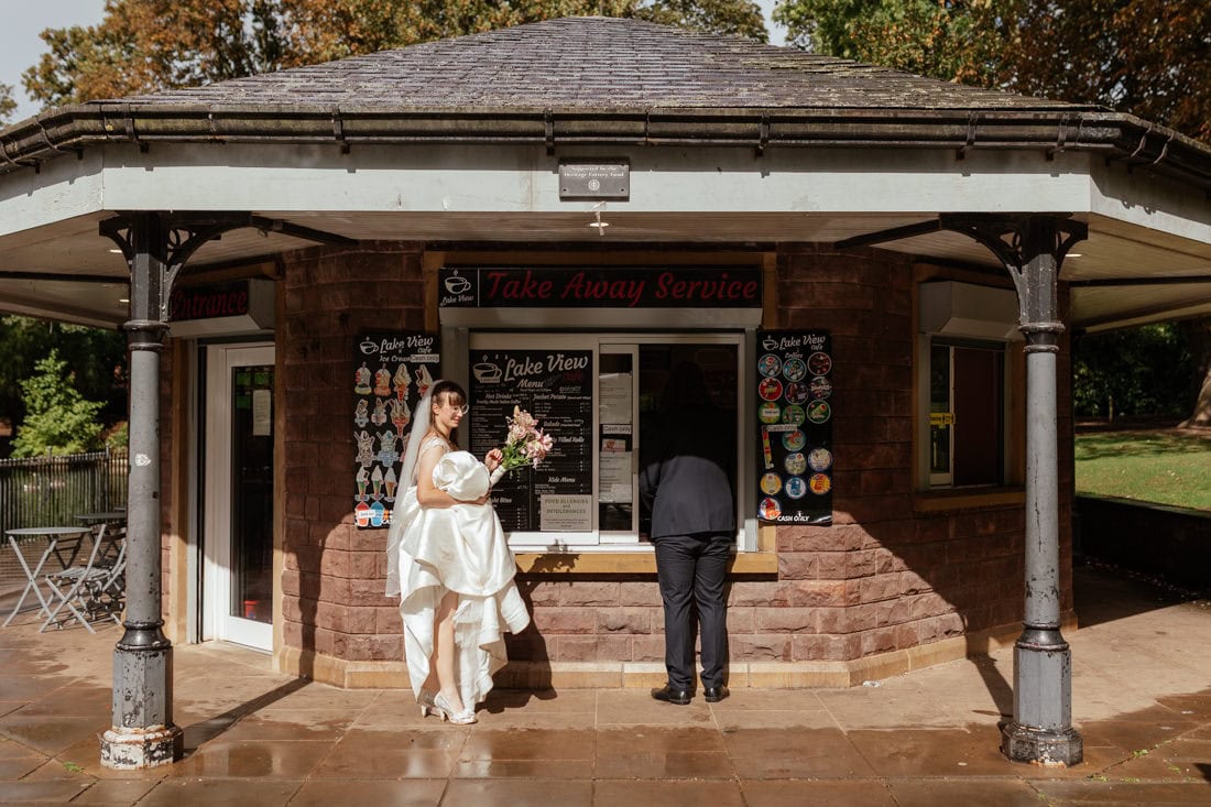 Bride smiles with bouquet as partner orders Ice-cream at kiosk, surrounded by rain-soaked pavement.