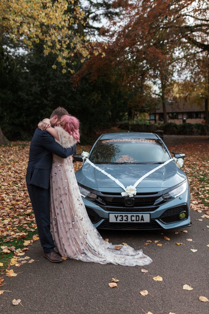 bride and groom embrace she's wearing a pink gown they're stood in front of a grey wedding car in an park surrounded by fallen leaves