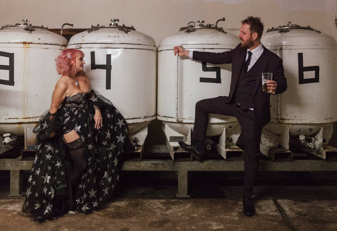 Two people in wedding attire pose playfully in front of large white brewing tanks labelled with numbers
