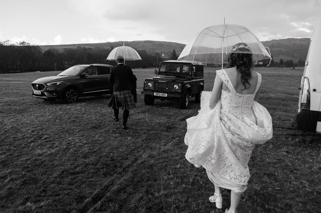 Bride and groom walking with clear umbrellas across a rain-soaked Scottish field towards a Land Rover Defender, in black and white.