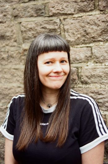 person smiling and looking into the camera stood in front of a stone wall