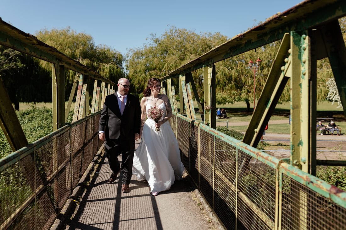 Couple walk across a narrow metal bridge, one in a wedding dress holding a bouquet and the other in a suit.