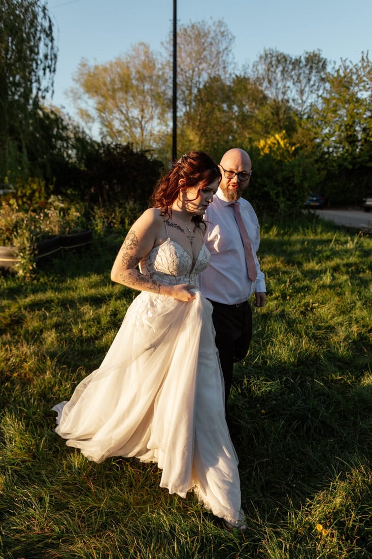 Couple walk across sunlit grass, one in a lace wedding gown and the other in a shirt and tie.
