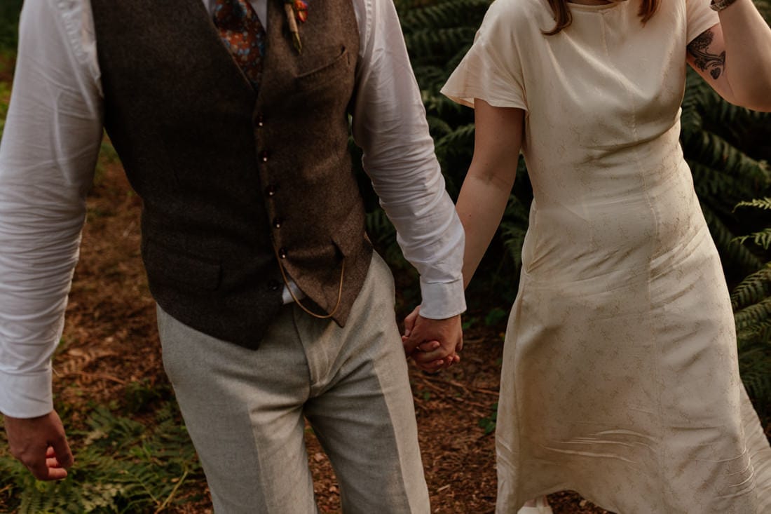 Couple holding hands while walking through forest, dressed in wedding attire with ferns and dirt path in background.