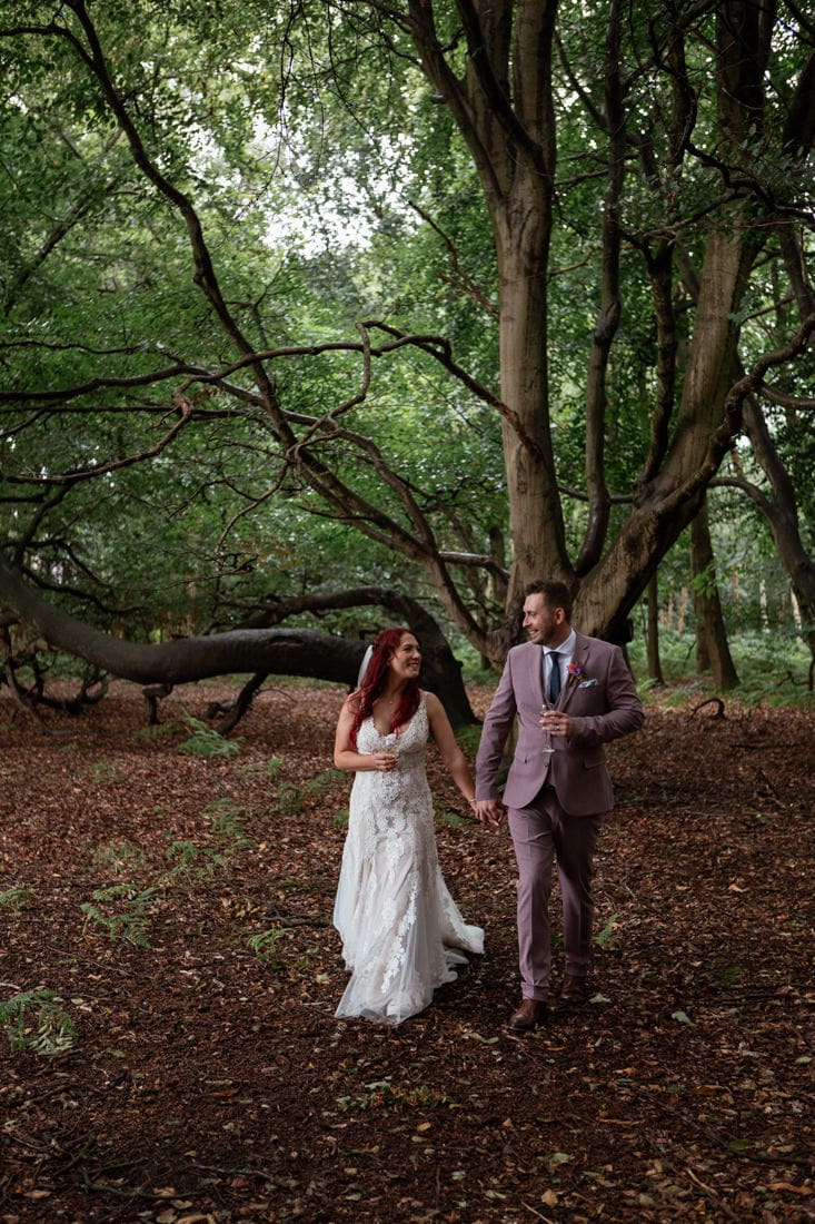 Bride and groom walking hand in hand through Sherwood Forest, beneath a sweeping ancient tree on a leaf-covered woodland floor.