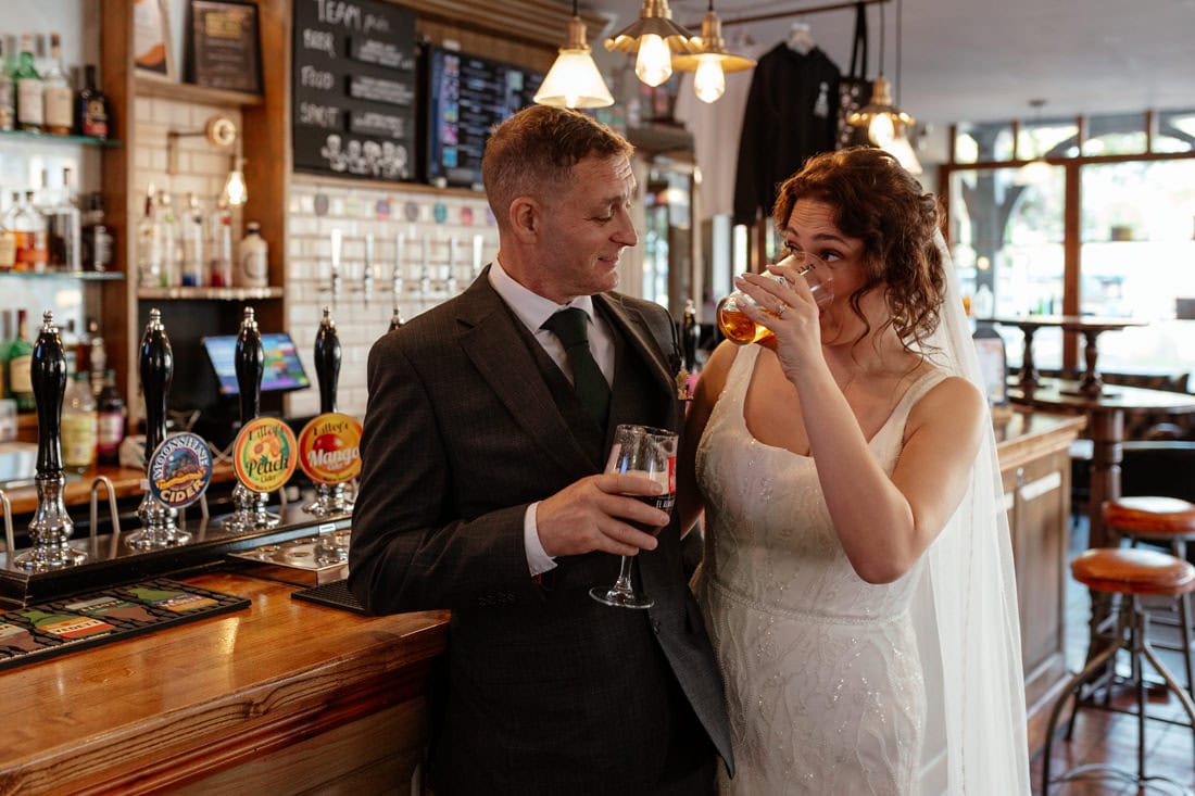 Bride and Groom share drinks at a wooden bar, chatting and smiling, she's drinking a pint