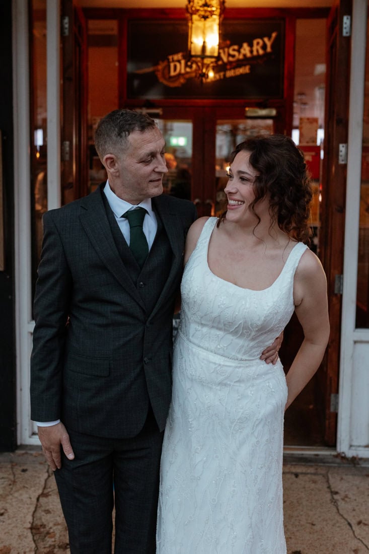 Couple stand smiling looking at each other outside The Embankment's Dispensary pub, dressed in wedding attire